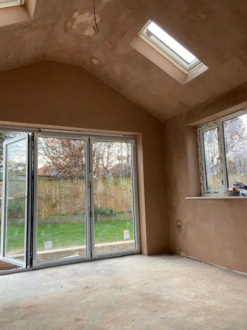 A freshly plastered room with large glass doors, a window, and a skylight, overlooking a grassy backyard with a wooden fence and some trees. The floor is bare, suggesting recent construction or renovation.