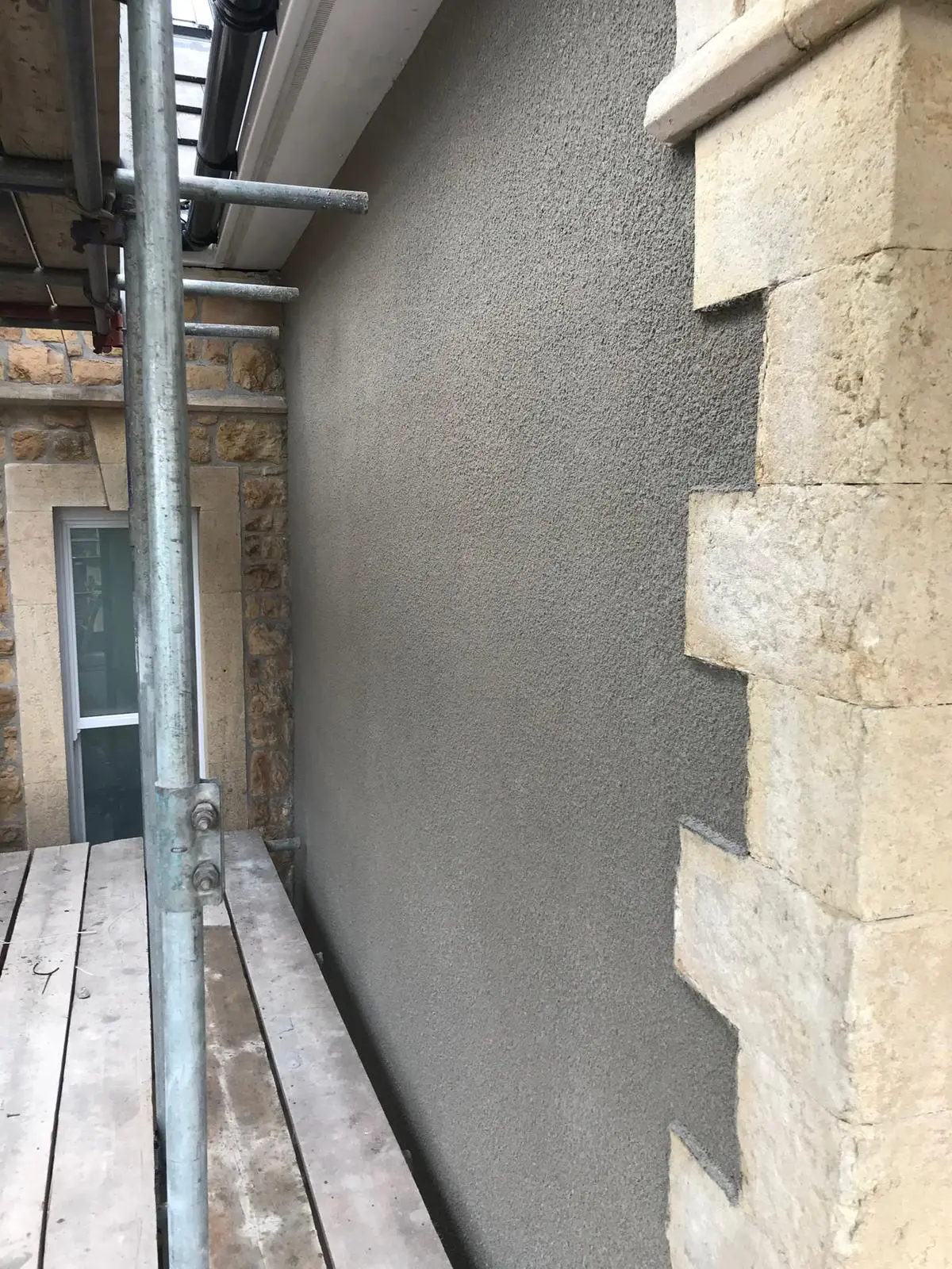 A freshly rendered exterior wall next to a stone corner of a building, with scaffolding and wooden planks in the foreground.