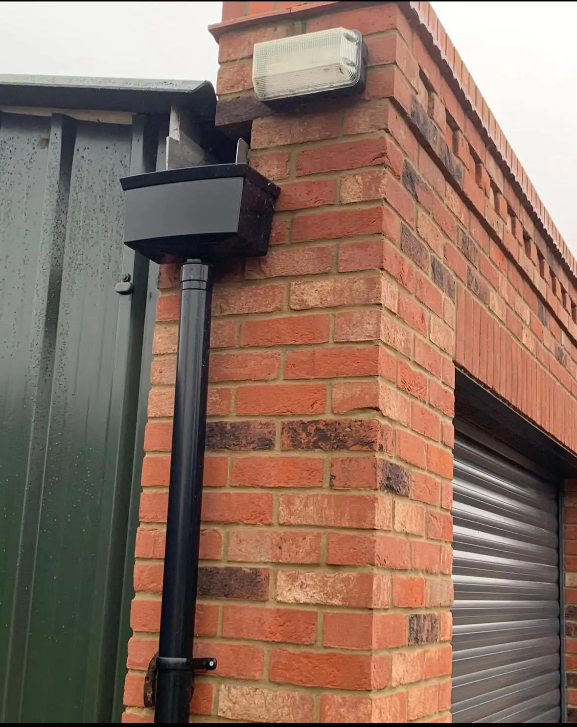 A black security camera is mounted on a red brick wall near a metal garage door and an outdoor light fixture. The camera is attached to a black pipe running vertically down the wall.