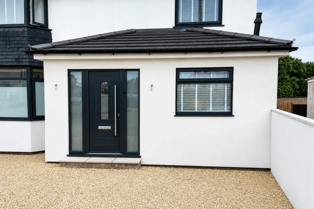 A modern white house with a black front door and a window with white blinds. The entrance has two narrow glass panels on either side of the door and a gravel front yard.